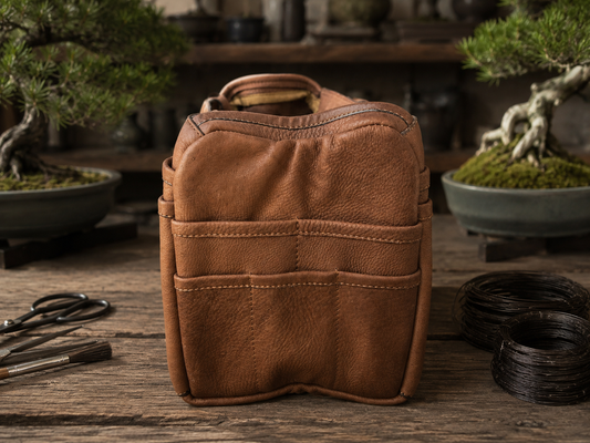 A brown leather Bonsai bag with top handles and front pockets, standing on a rustic wooden surface. Bonsai trees and tools are blurred in the background, creating a styled workshop-like setting.