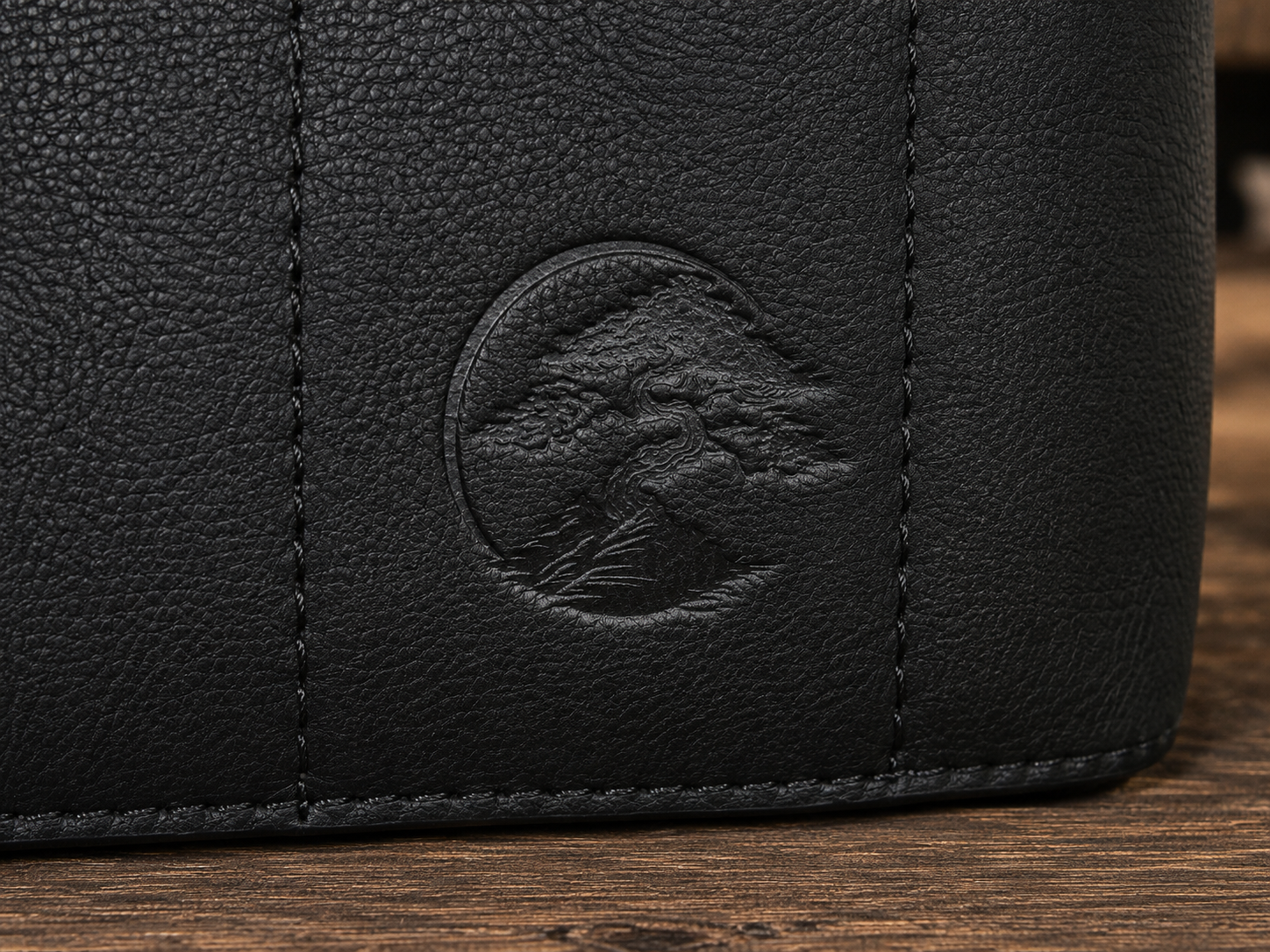 Close-up of a black Bonsai bag, showing a circular embossed logo of a bonsai tree on a wooden surface.