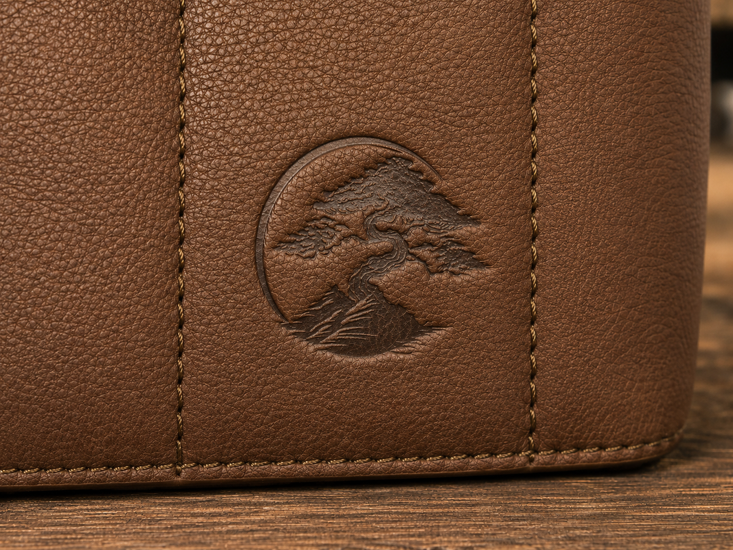 Close-up of the brown textured leather Bonsai bag, featuring a circular embossed logo of a bonsai tree on the front panel.