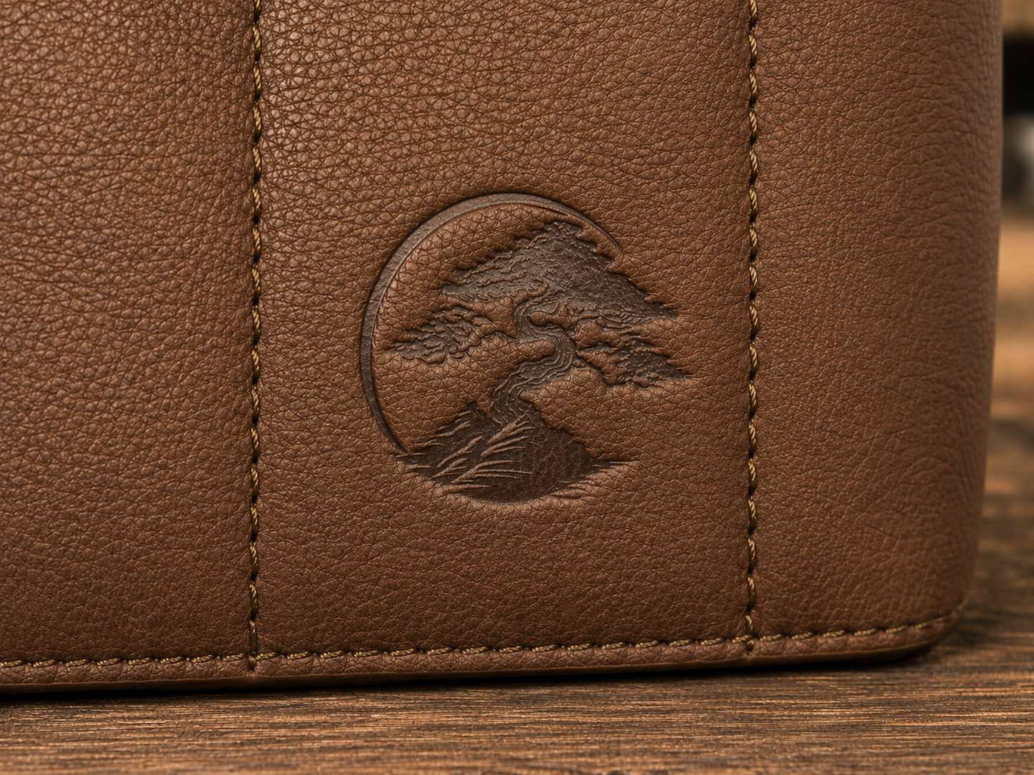 Close-up of the brown textured leather Bonsai bag, featuring a circular embossed logo of a bonsai tree on the front panel.