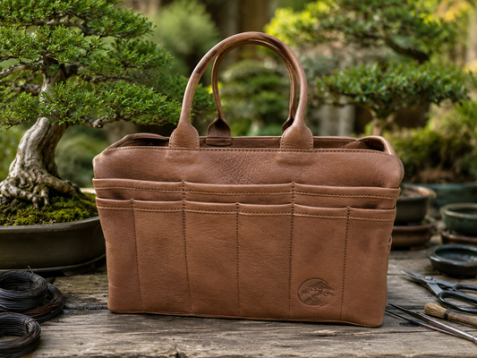 A brown leather Bonsai tote bag with dual top handles and multiple front pockets, displayed on a rustic wooden surface with bonsai trees and gardening tools in the background.