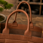 A close-up of a brown leather Bonsai bag with dual top handles, set against a warm, blurred background featuring a wooden surface and a small bonsai tree.