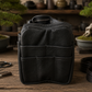 A black leather Bonsai bag with multiple front pockets, displayed on a wooden table with bonsai trees and crafting tools in the background.