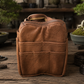A brown leather Bonsai bag with top handles and front pockets, standing on a rustic wooden surface. Bonsai trees and tools are blurred in the background, creating a styled workshop-like setting.