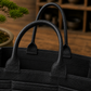 Close-up of a black pebbled leather Bonsai bag with dual top handles, set against a warm wooden background with a blurred bonsai tree.