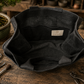 A black leather Bonsai bag, shown from a top-down angle on a rustic wooden workbench surrounded by Bonsai-tools and a bonsai tree. he inside highlights four large pockets and the SECHI's logo. 
