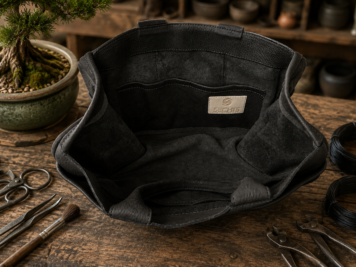A black leather Bonsai bag, shown from a top-down angle on a rustic wooden workbench surrounded by Bonsai-tools and a bonsai tree. he inside highlights four large pockets and the SECHI's logo. 
